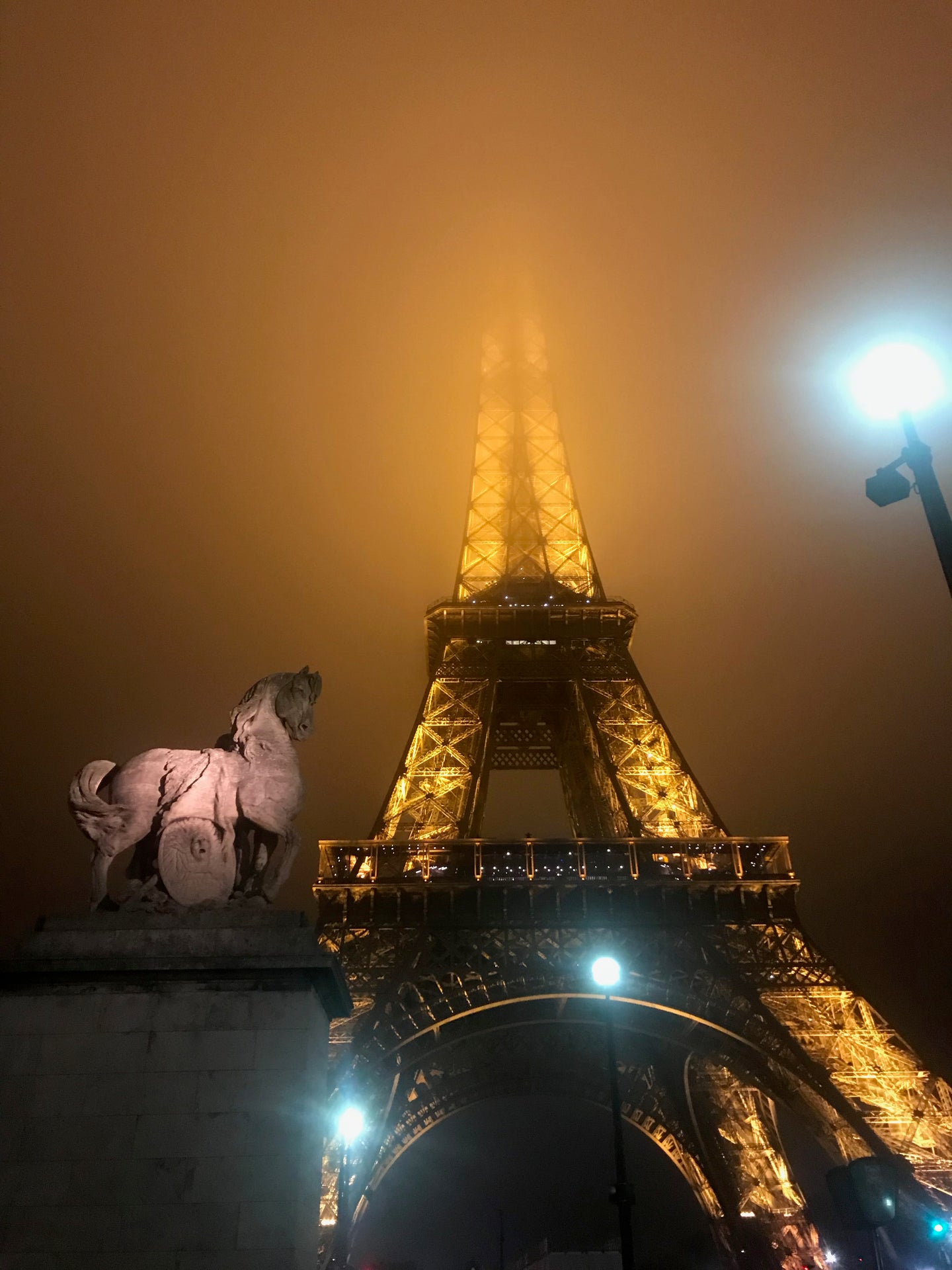picture of the eiffel tower at night lit up against a background of fog, a street lamp and statue of a horse in the foreground